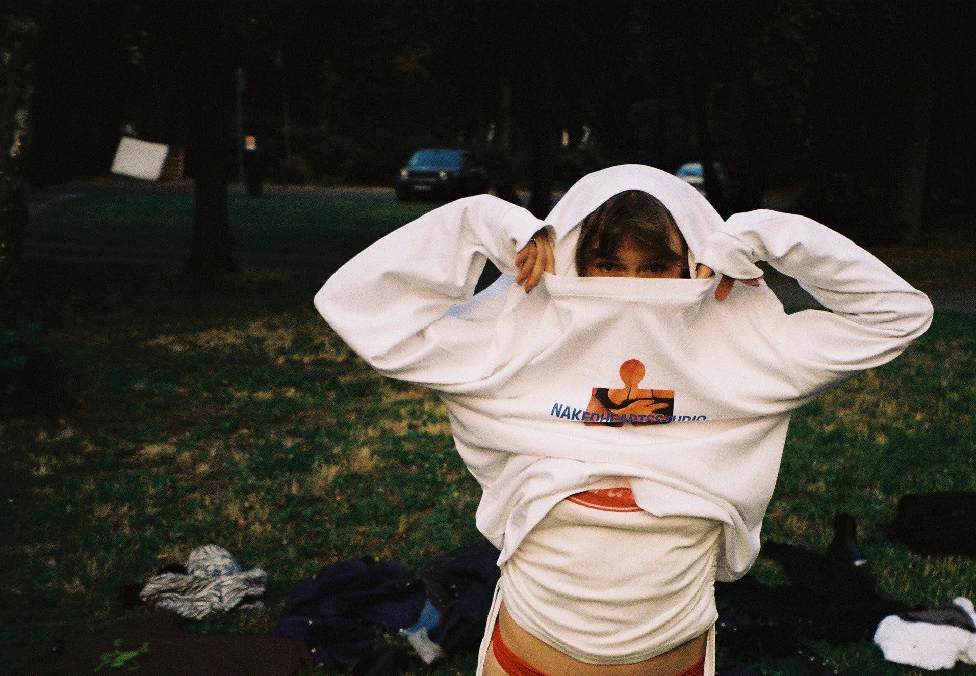 A person standing on grass outdoors, wearing a white oversized hoodie with a graphic print on the chest and red underwear. Clothes are scattered on the grass around them, and trees and a dark background are visible.