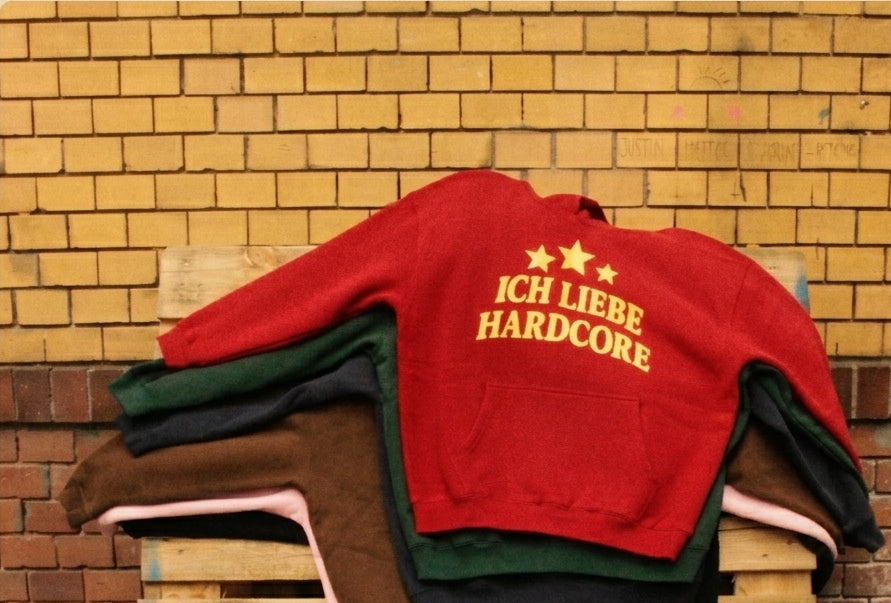 A stack of sweatshirts in red, green, brown, and pink, with the top red sweatshirt featuring yellow text that reads 'ICH LIEBE HARDCORE' and two yellow stars, displayed on a wooden pallet against a brick wall.